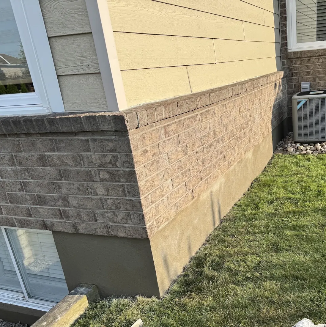 Parged foundation corner beneath tan siding and brick with a basement window